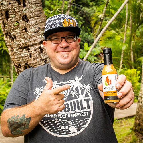 Smiling man holding a hot sauce bottle in a tropical setting.