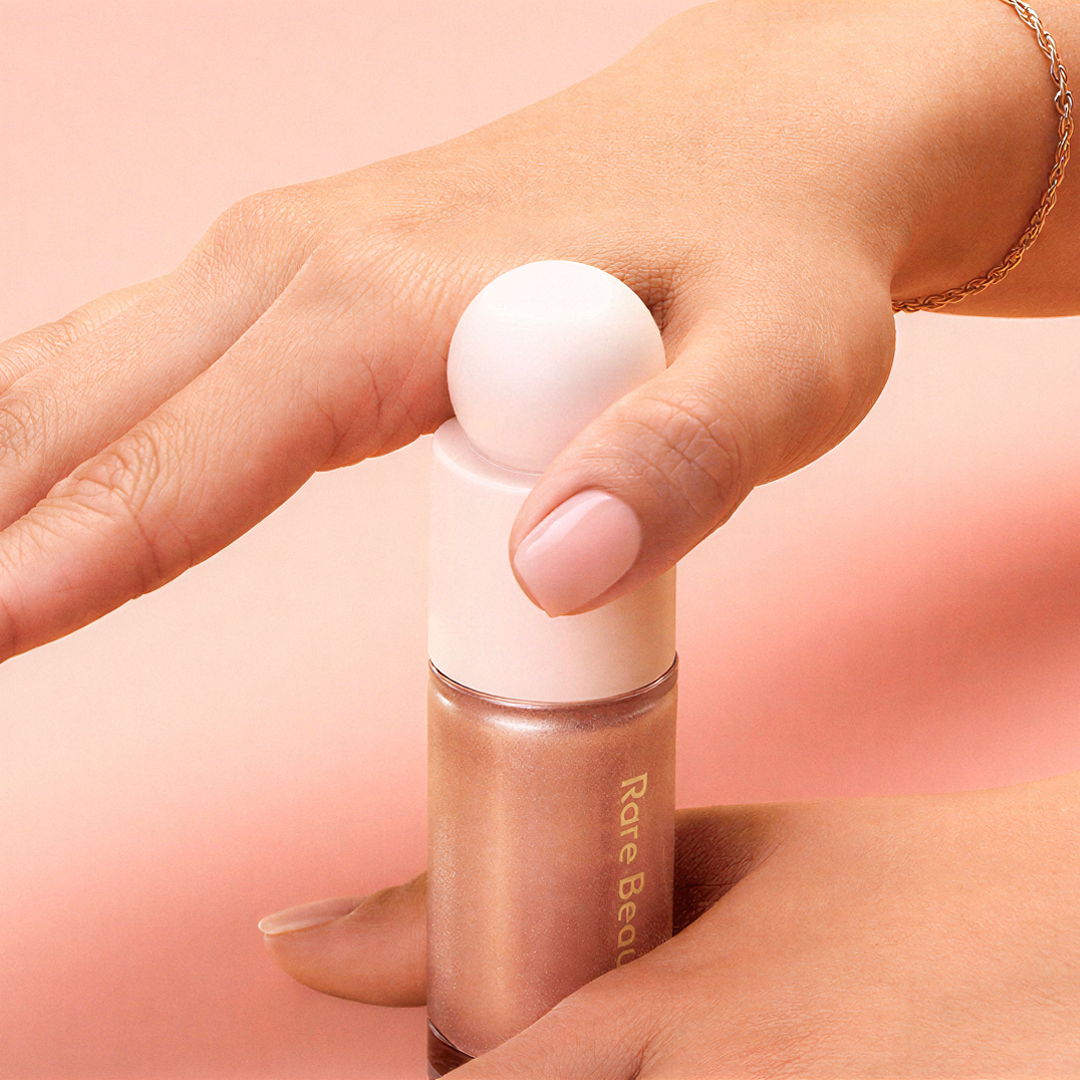 Hand holding a makeup bottle with a white cap on a pink background.