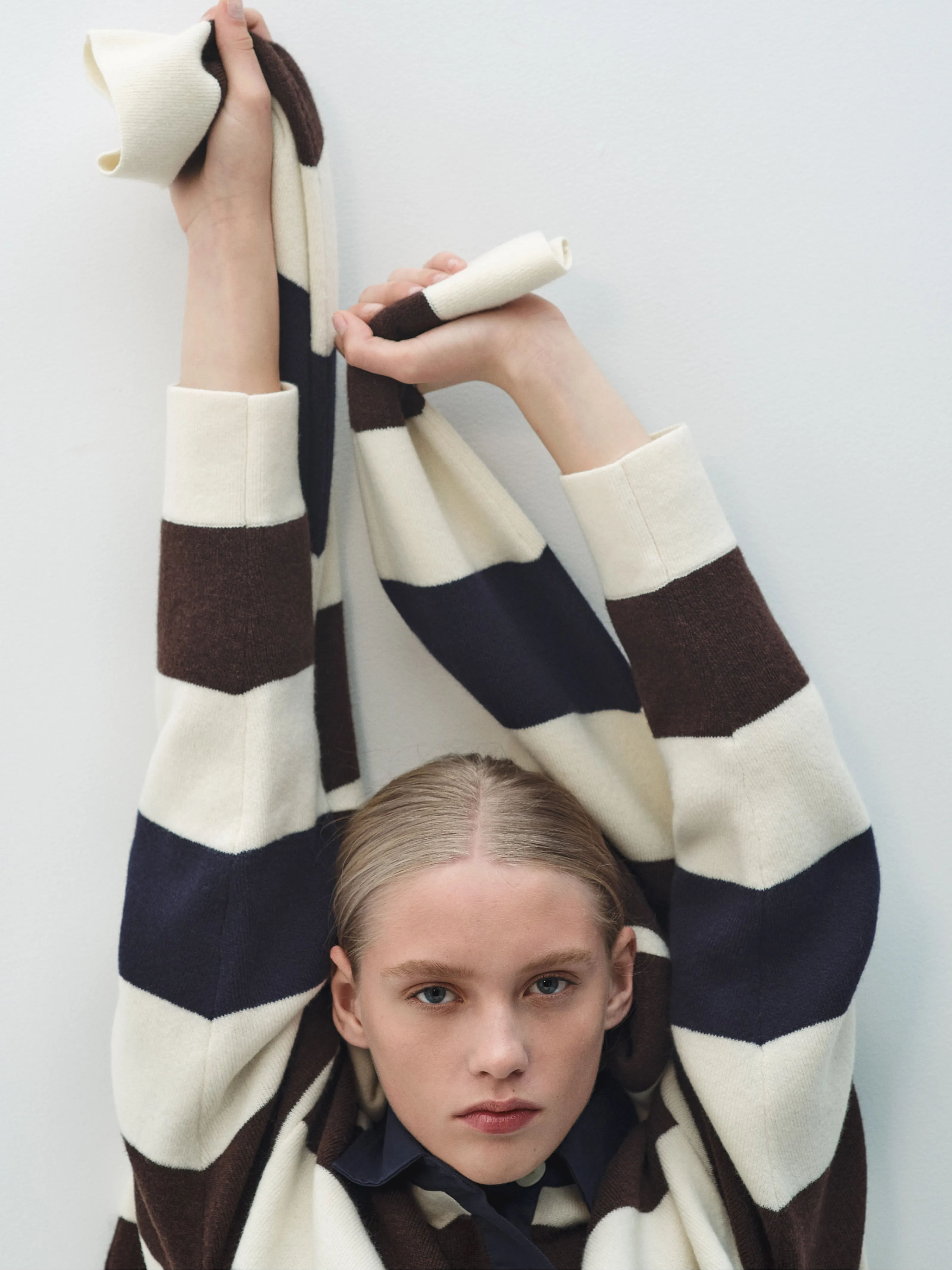 A model poses with arms raised, wearing a striped sweater.