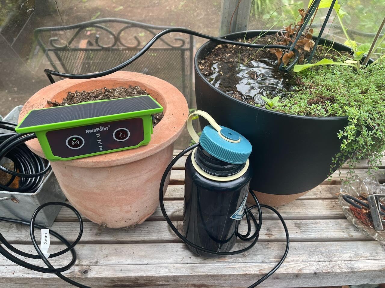 Potted plants with a watering device and electronic equipment on a wooden surface.