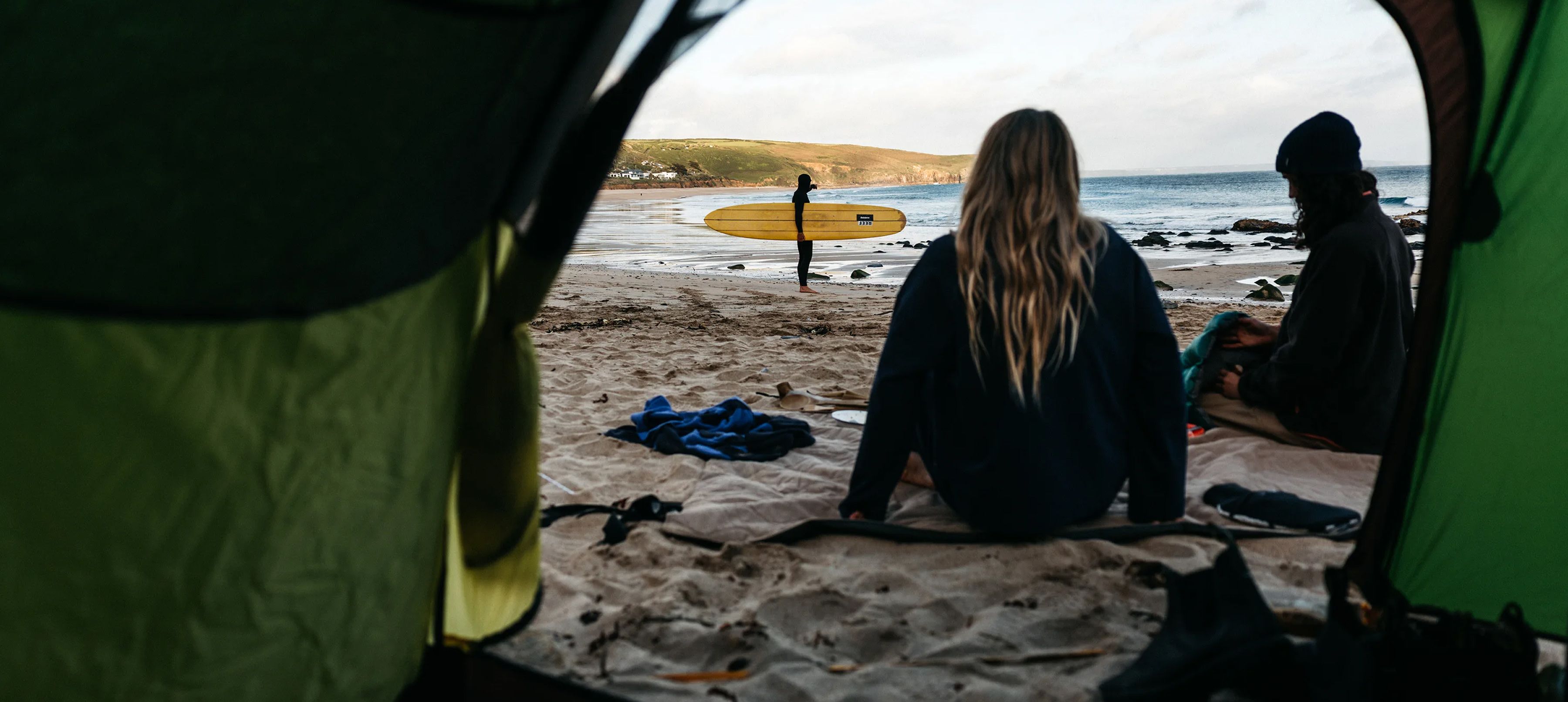 Two people sitting in a tent, looking at the beach and surfer.