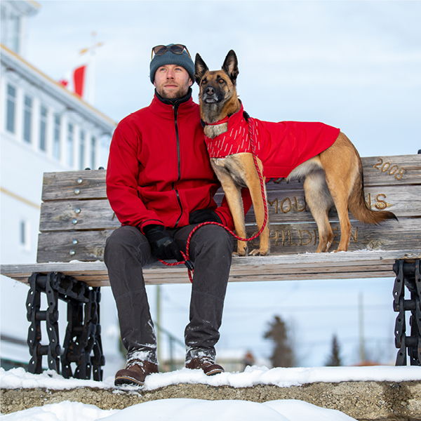 Man in red coat sitting on bench with a dog in winter setting.