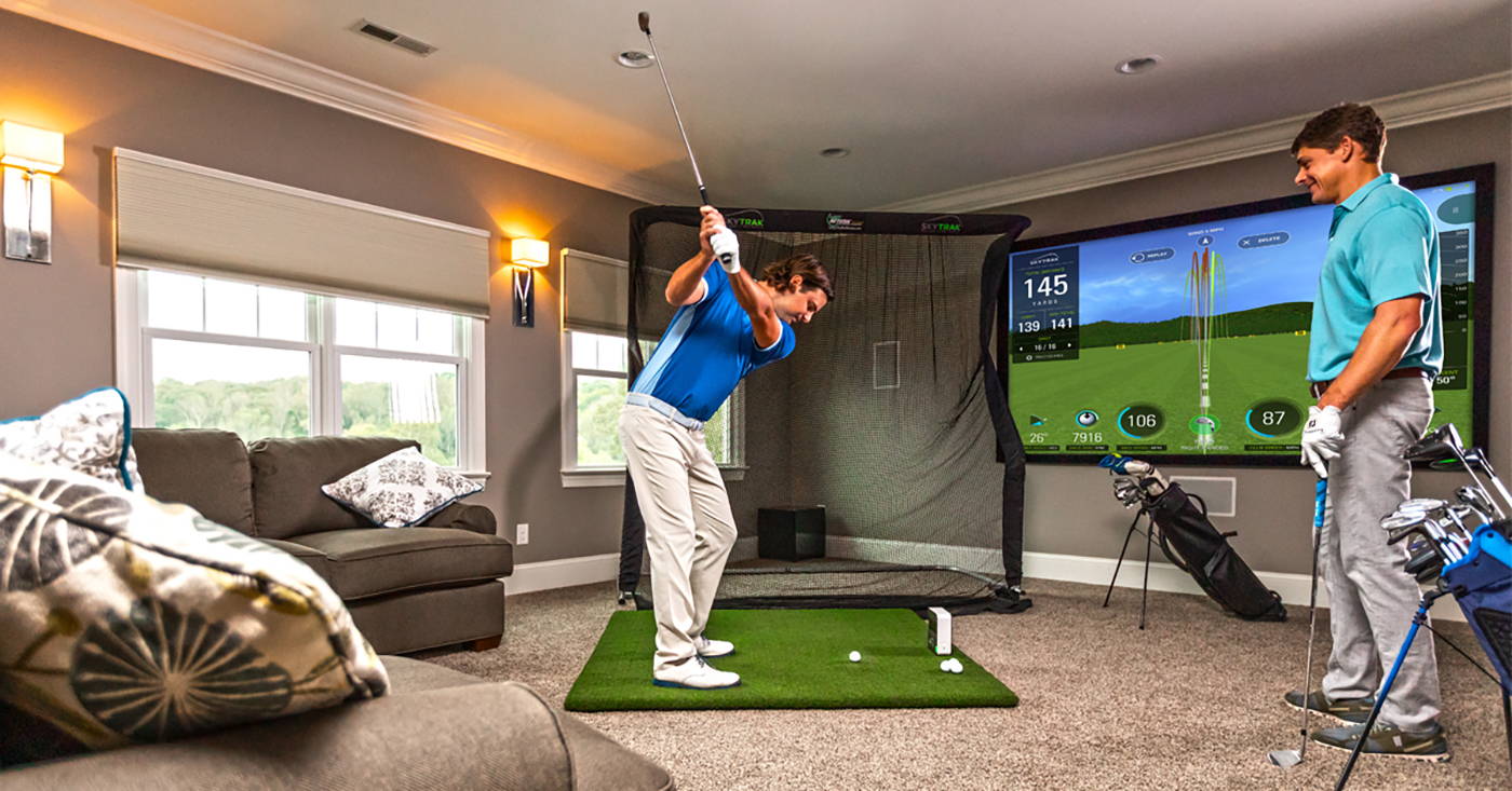 One man swinging into a net in a living room using a SkyTrak golf simulator while another man watches