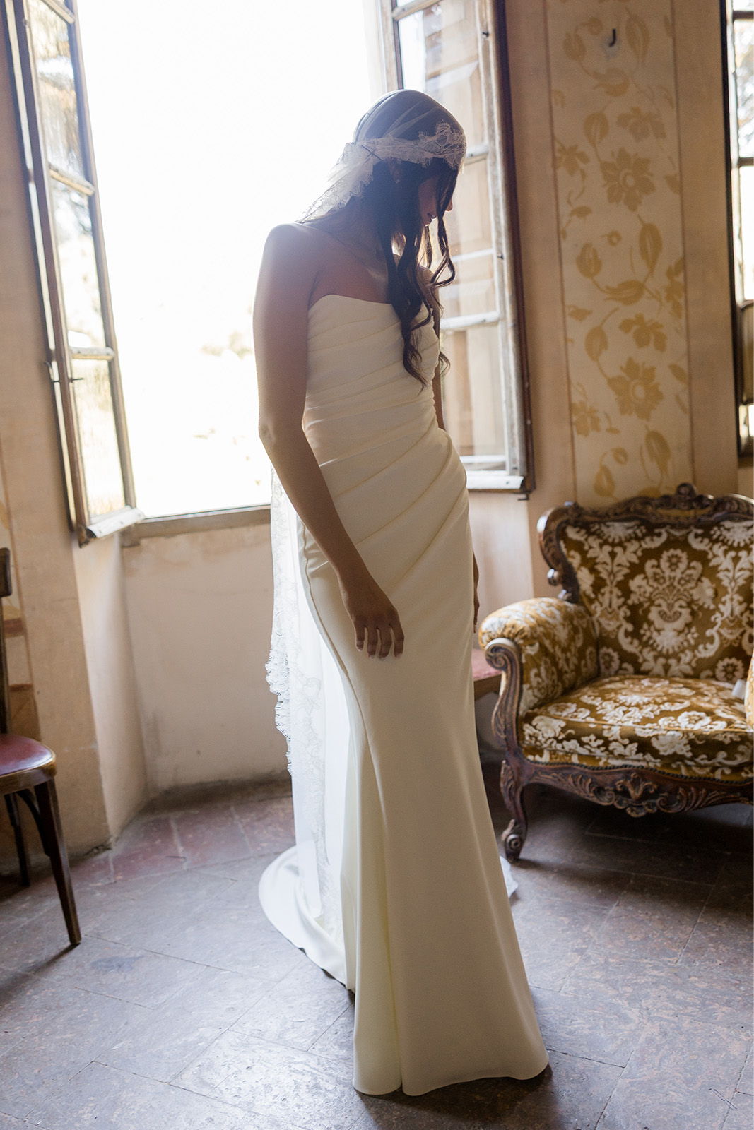 Bride in a white dress standing near an open window.