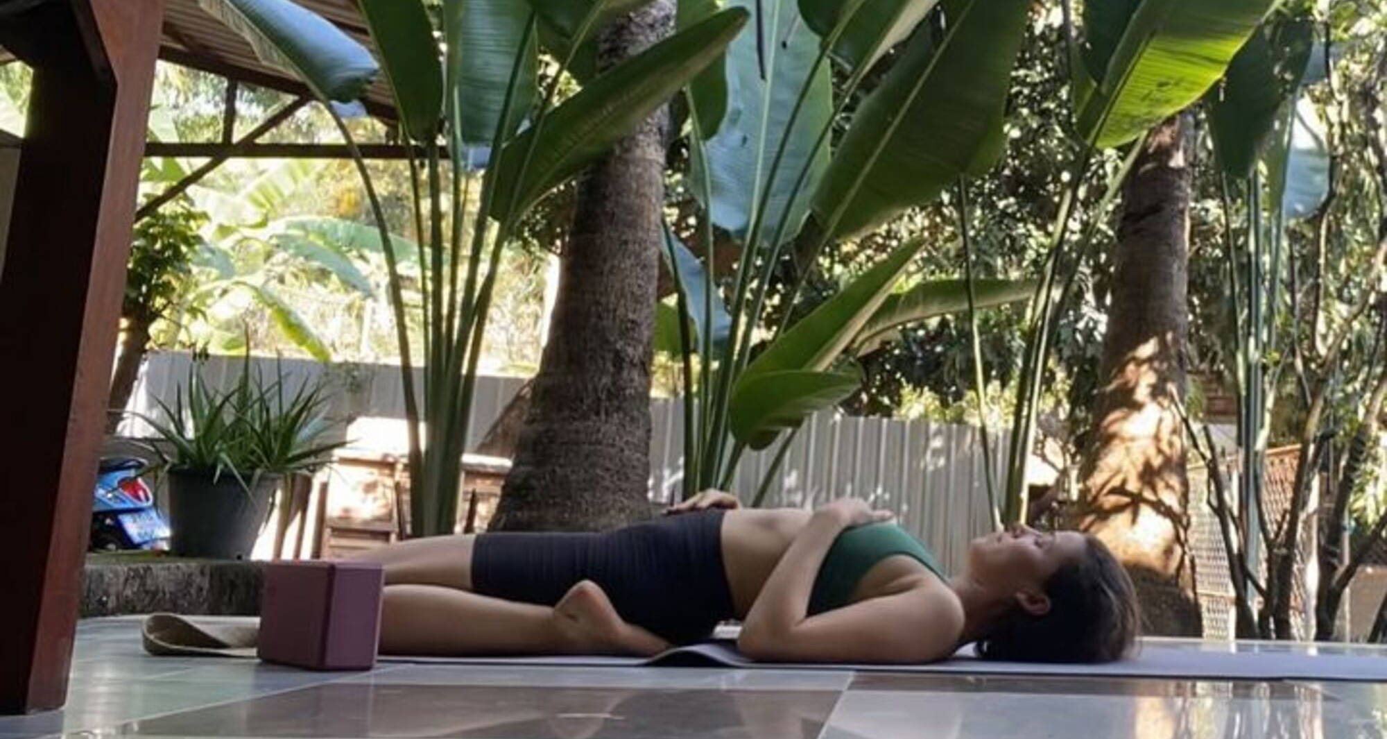 woman in a green bra and black bike shorts lays down in a yoga pose surrounded by greenery