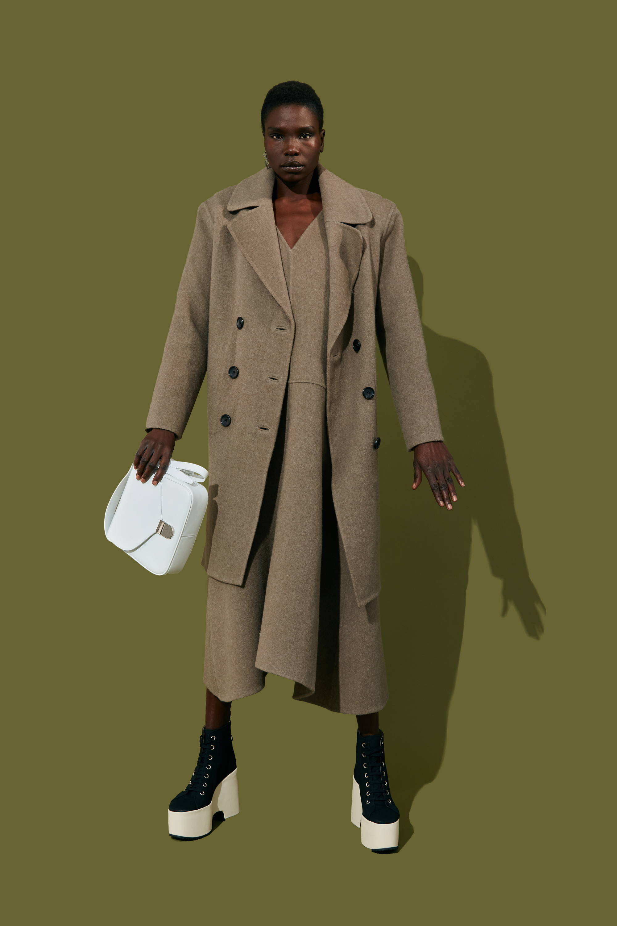 Model wearing a stylish beige coat and platform boots, holding a white handbag.
