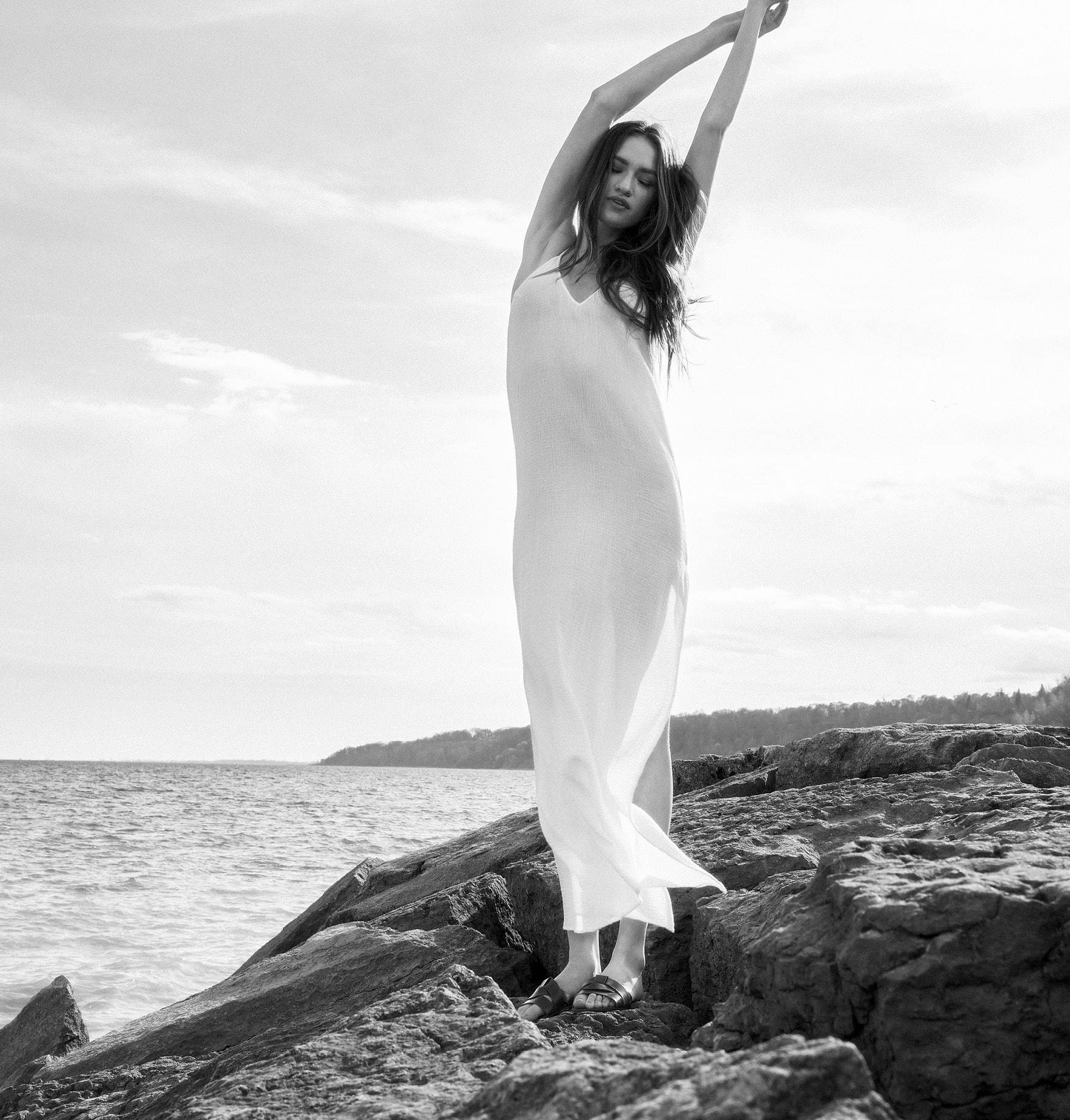 Tall woman standing on rocks with her hands in the air wearing a white gauze cover up dresss