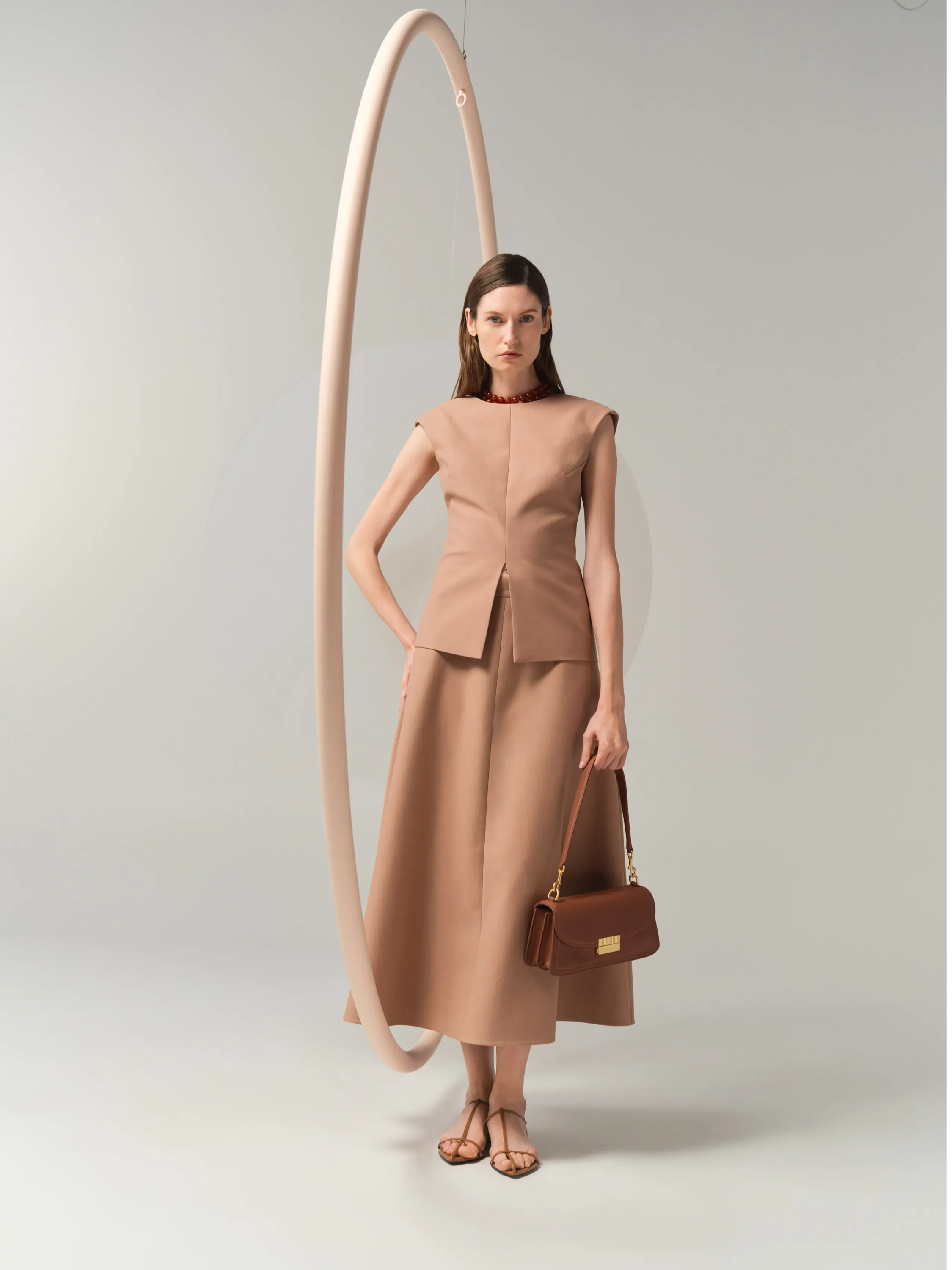 Model wearing a beige outfit with a handbag, standing near a circular backdrop.
