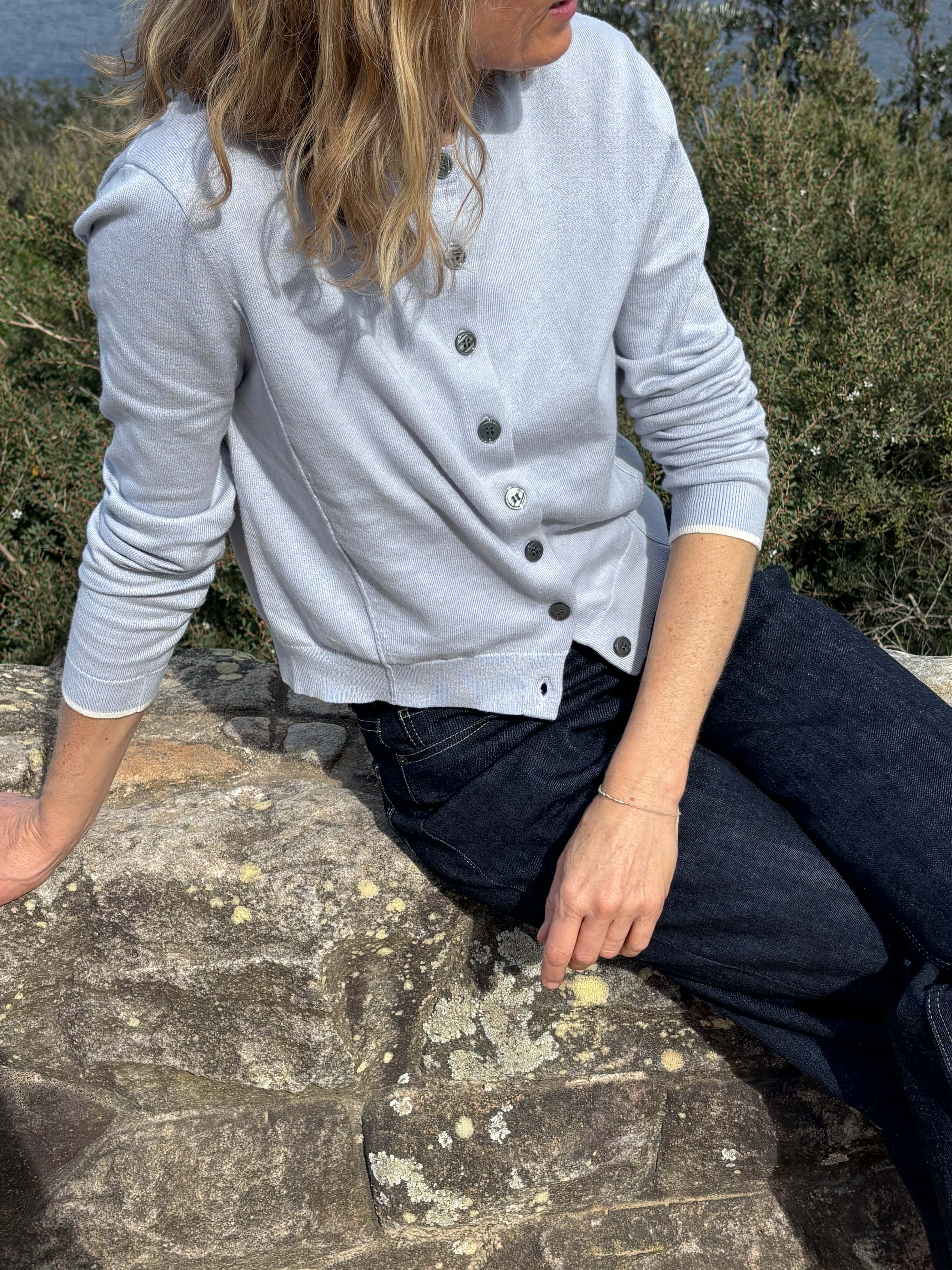 Person sitting on a stone wall, wearing a light blue cardigan and dark jeans.