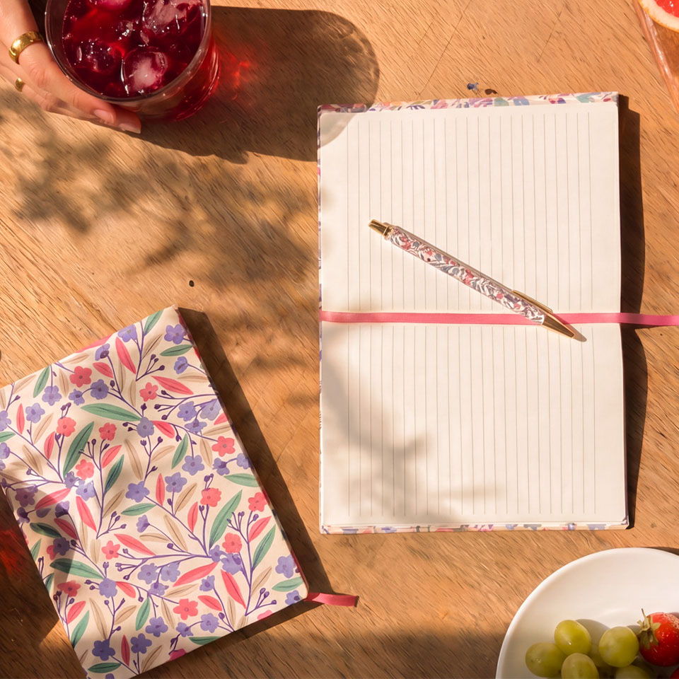 Two notebooks and a pen on a wooden table with a drink and fruit.