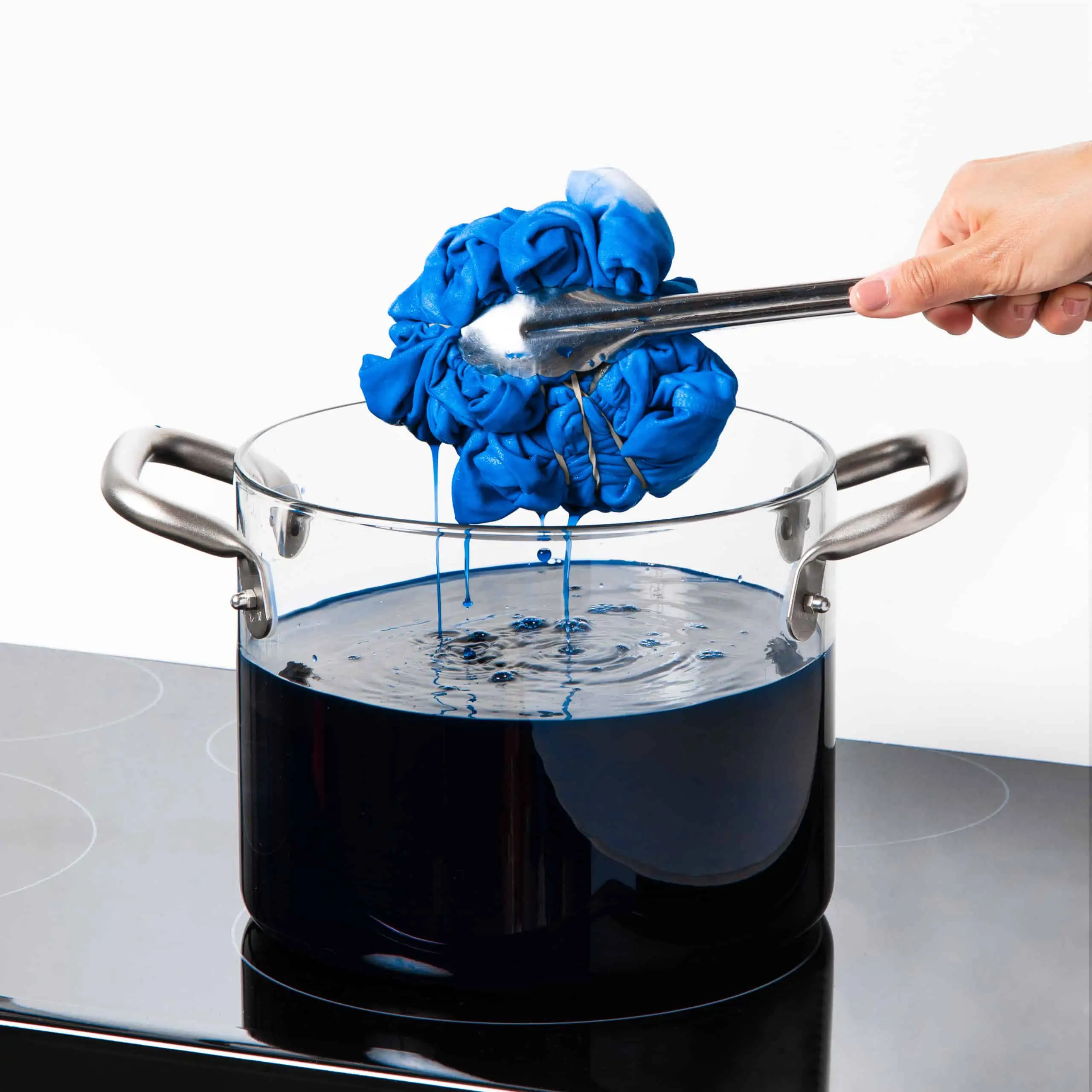 A hand using tongs to lift blue fabric from a pot of blue dye.