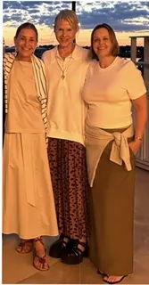 Three women standing together, smiling, wearing casual outfits, during sunset.