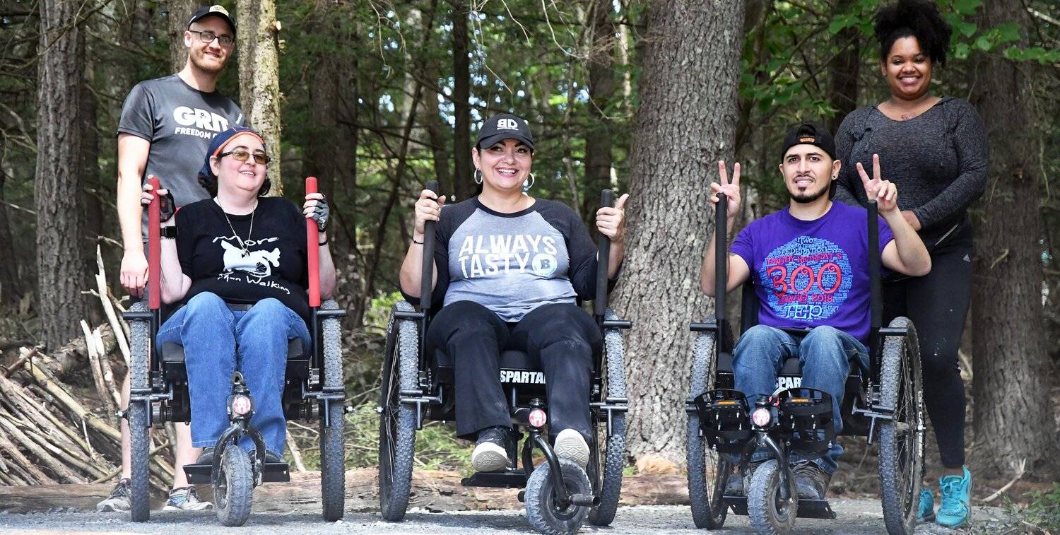 3 people in GRIT Freedom Chair all-terrain wheelchairs and 2 people standing behind them smile in front of wooded tree area