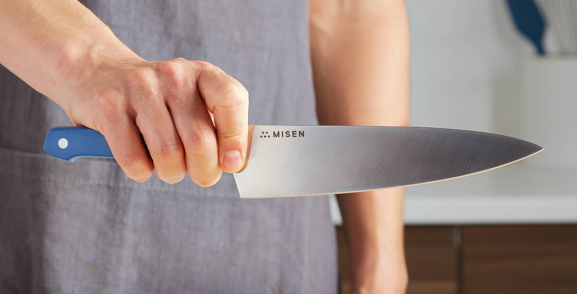 A chef holds a blue Misen Chef’s Knife towards the camera, with the blade’s steel and Misen logo clearly visible.