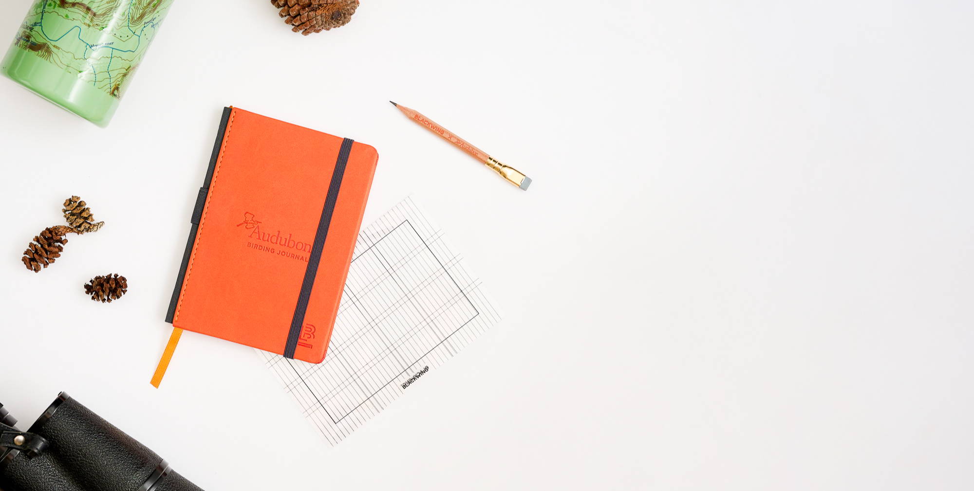 Flat lay of the red Audubon Journal with the template and pencil next to it. Binoculars, pinecones, and a green water bottle as props on a white background.