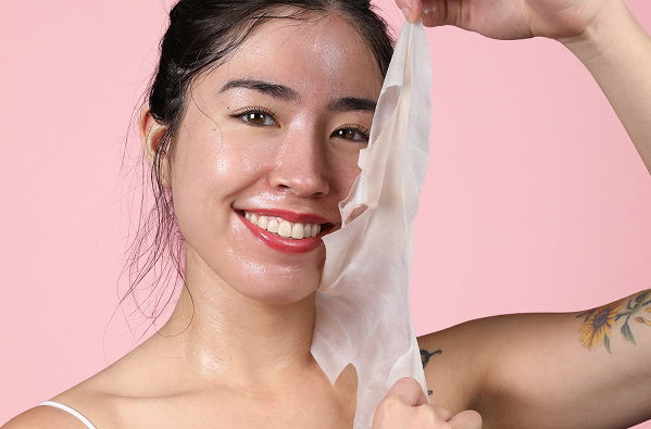 Smiling woman holding a skincare mask against a pink background.