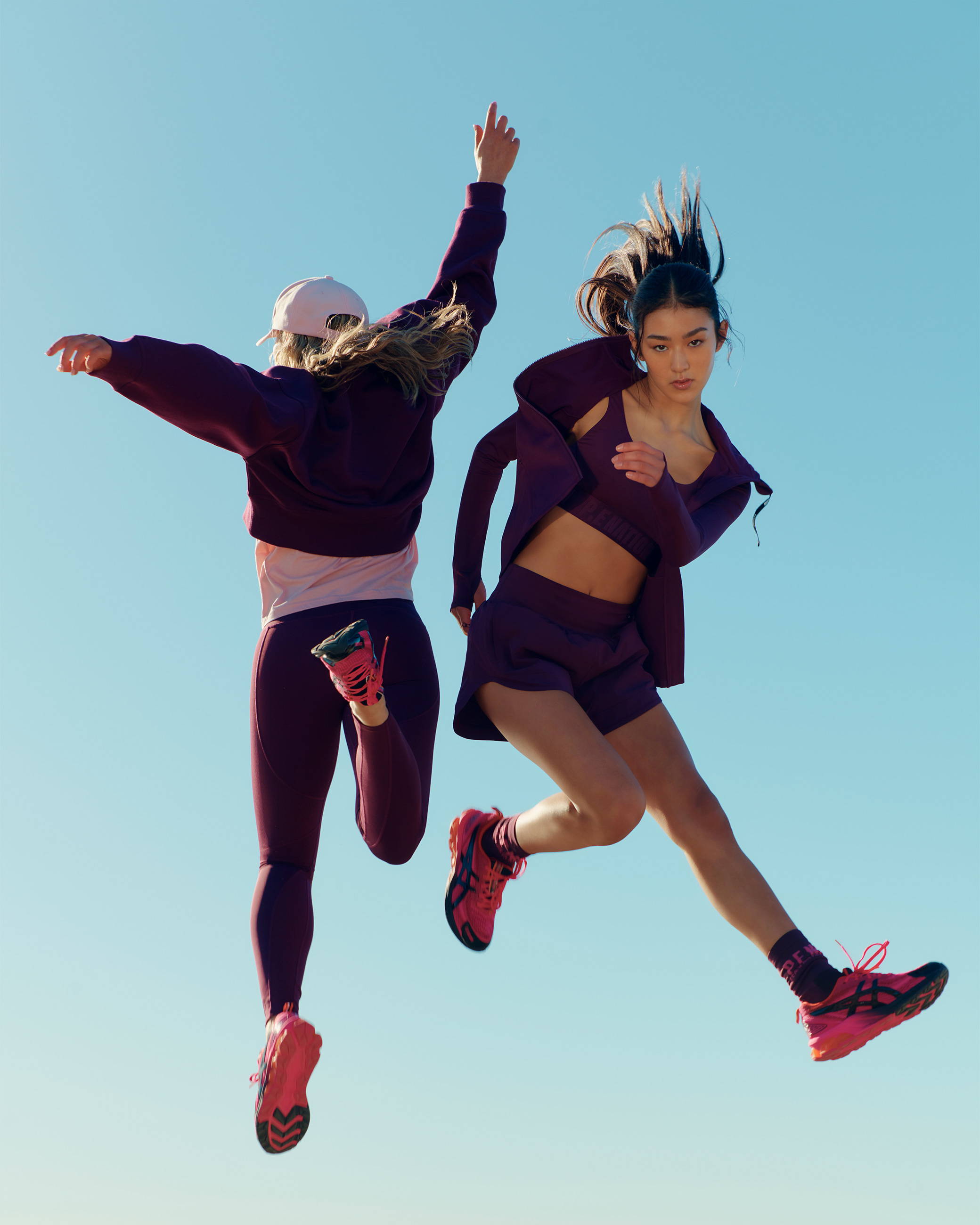 Two girls are jumping in the air wearing a range of hiking clothing. It is a cold but sunny day.