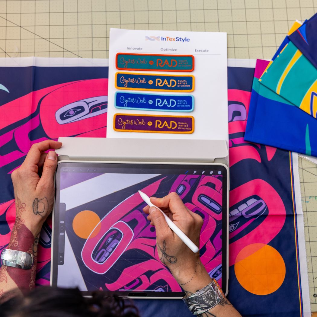 Artist Crystal Worl drawing on a tablet with colorful fabric swatches nearby.
