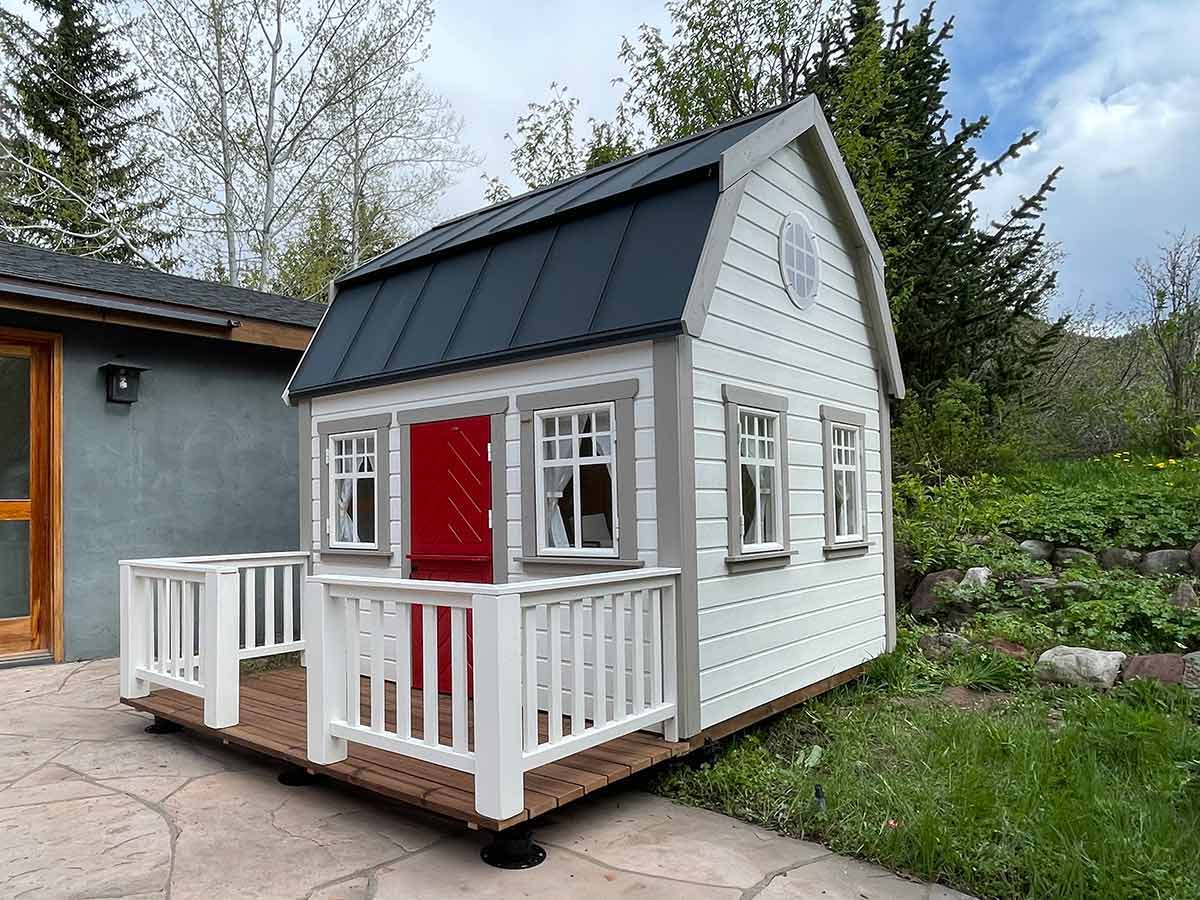 Kids Outdoor Playhouse Farmhouse with red dutch door, 4 opening windows and black metal roof in the backyard by WholeWoodPlayhouses