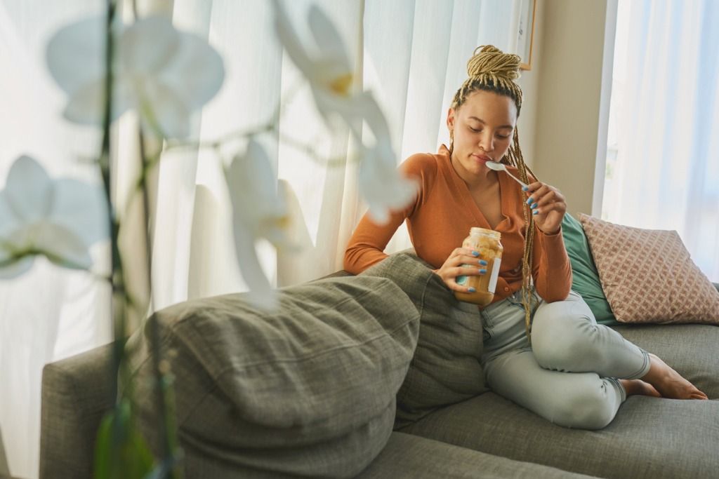 Woman eating ice cream on a couch, surrounded by soft cushions and plants.