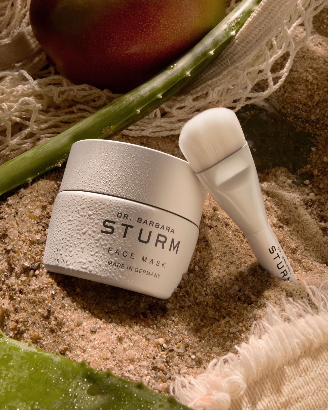 A skincare cream jar and applicator on a sandy surface with plants.