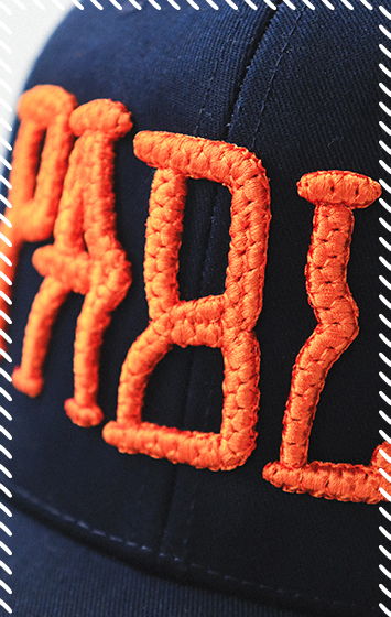 Close-up of navy cap with raised orange quilted puff embroidery of stylized lettering, showing the quilted texture created with stitching pattern