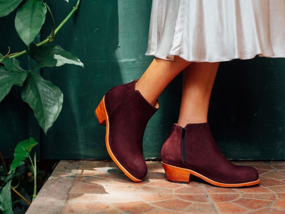 An outdoor lifestyle product shot from the calves down of a person wearing a flowy white dress or skirt standing on patterned terra-cotta floor tiles. The person is wearing deep burgundy or plum-colored suede ankle boots featuring a black side zipper and a low stacked wooden block heel, set against a dark teal or green painted wall with green tropical leaves framing the left side.