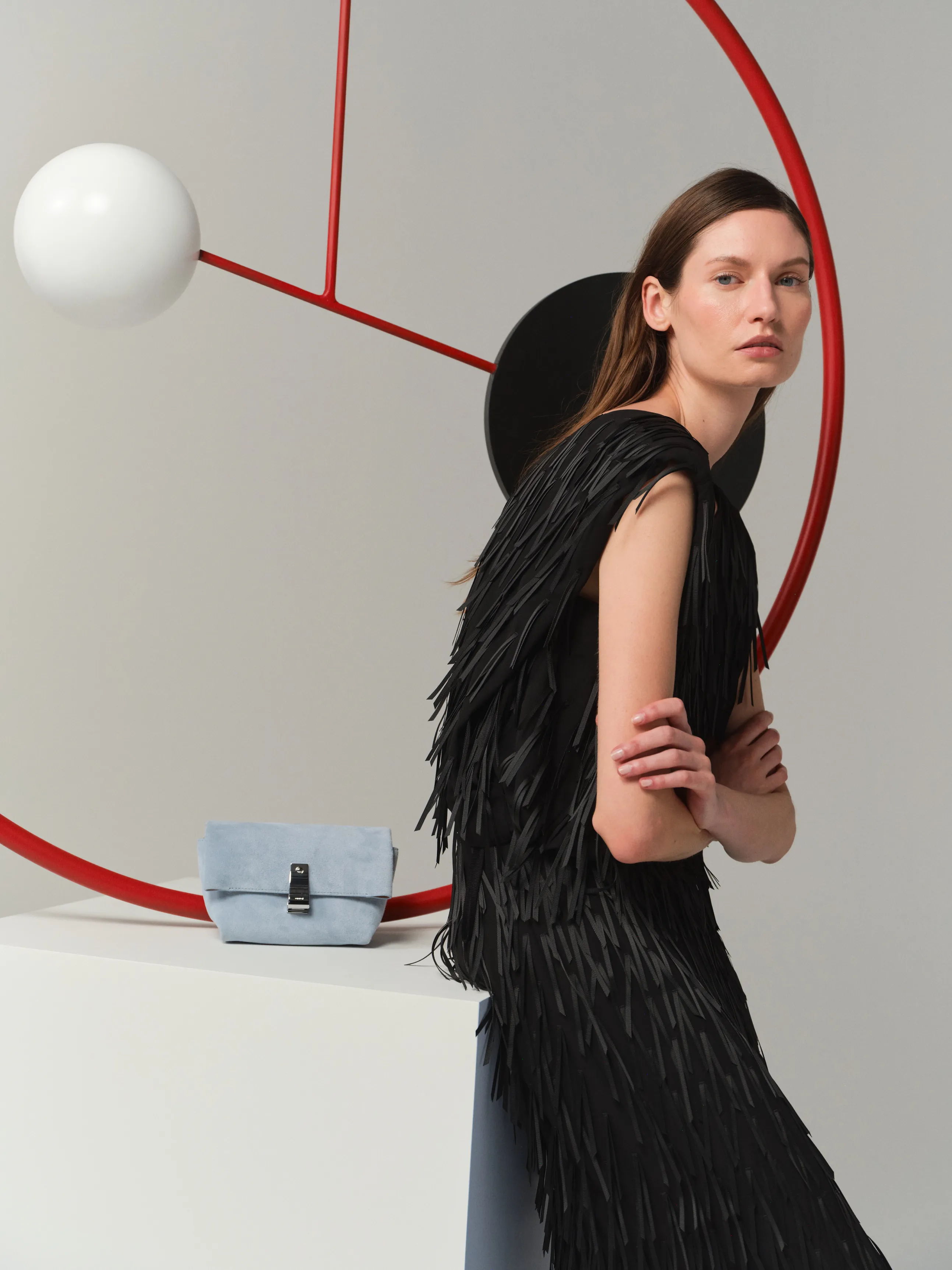 Model in a fringed black dress, posing beside a geometric backdrop and handbag.