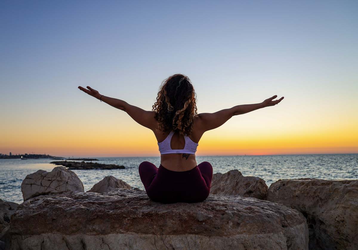 A woman facing a sunset and ocean sitting on rocks