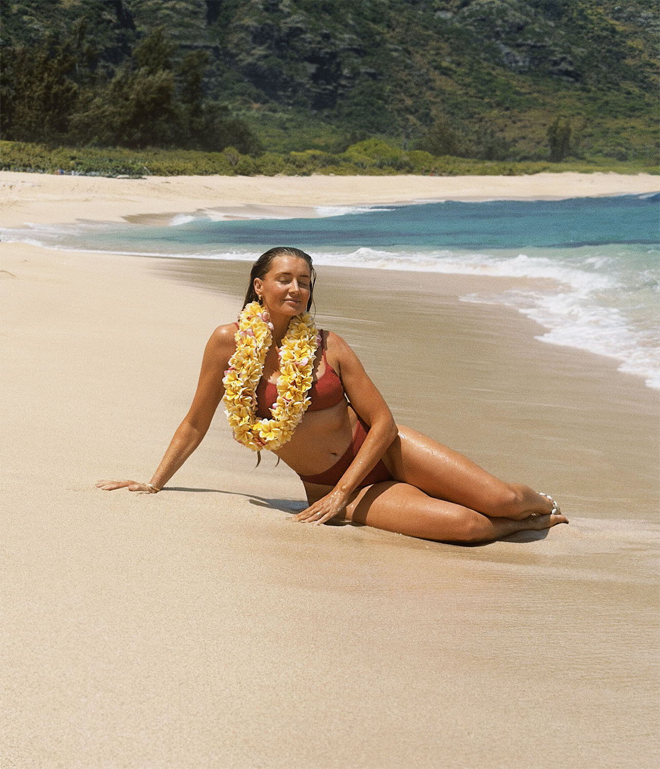 Woman in a bikini sitting on the beach, wearing a flower lei.