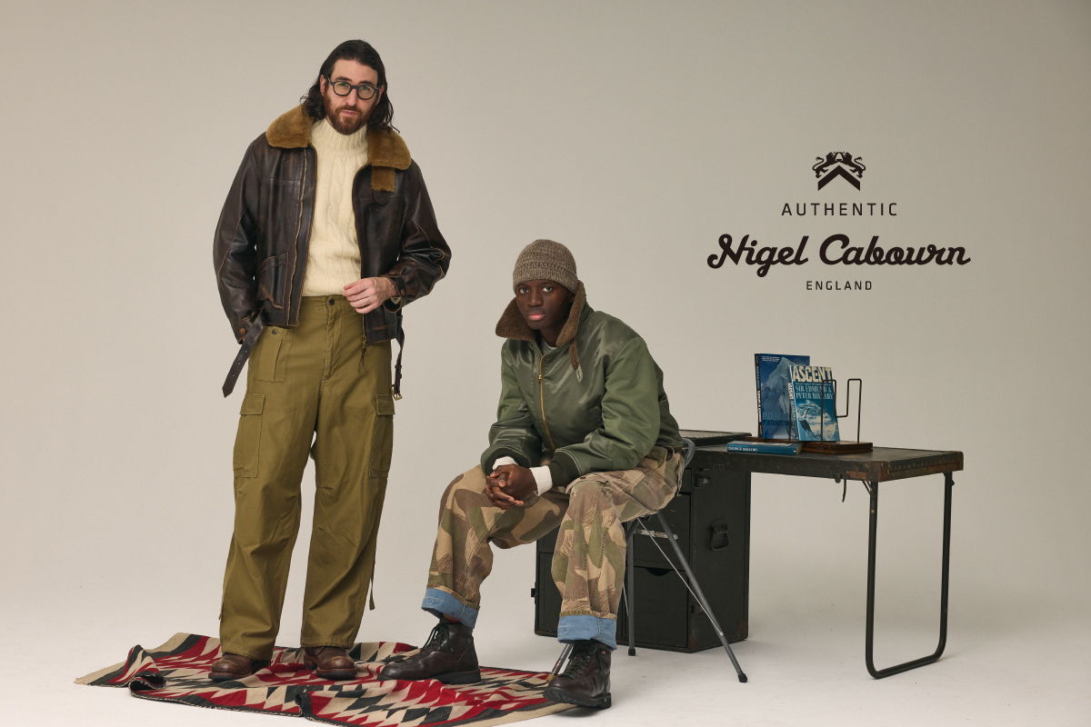 Two men in stylish outfits, one seated at a table, minimalistic background.