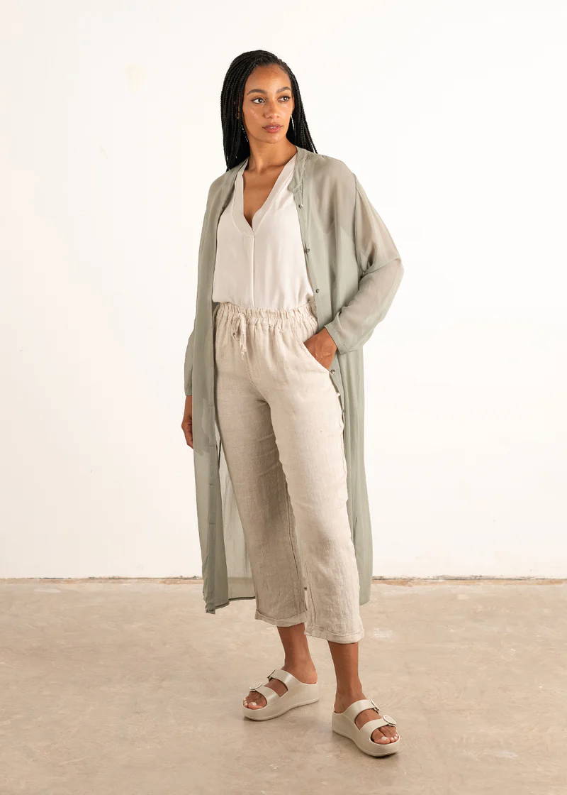 A model wearing a long, sage green semi sheer shirt over an off white top, linen trousers and chunky slides.
