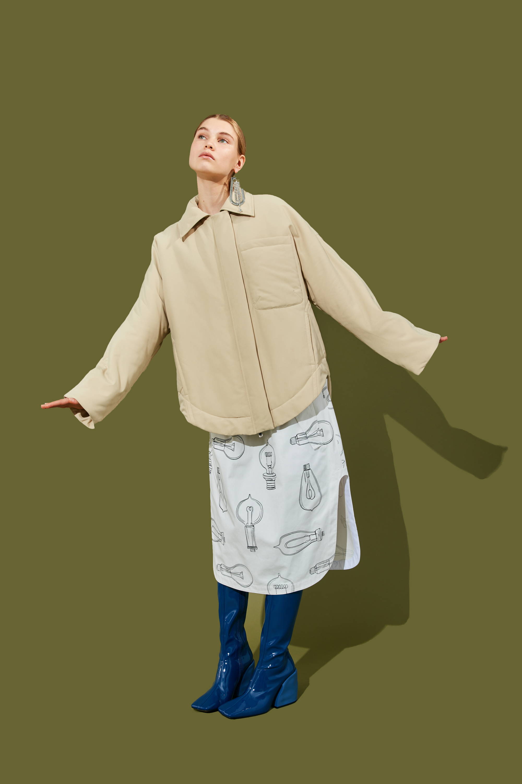 Model in oversized jacket and patterned skirt against a green background.