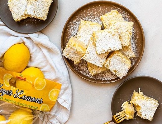 Lemon bars on plates with fresh lemons and a cloth.