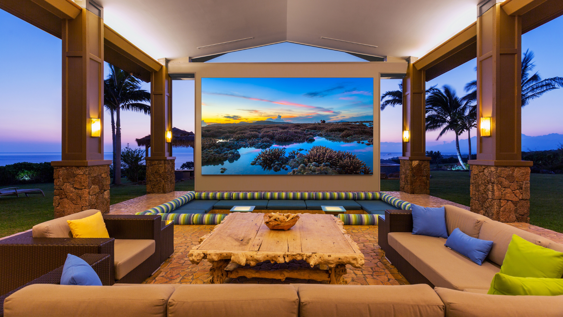 Outdoor living space with a large screen, colorful furniture, and sunset view.