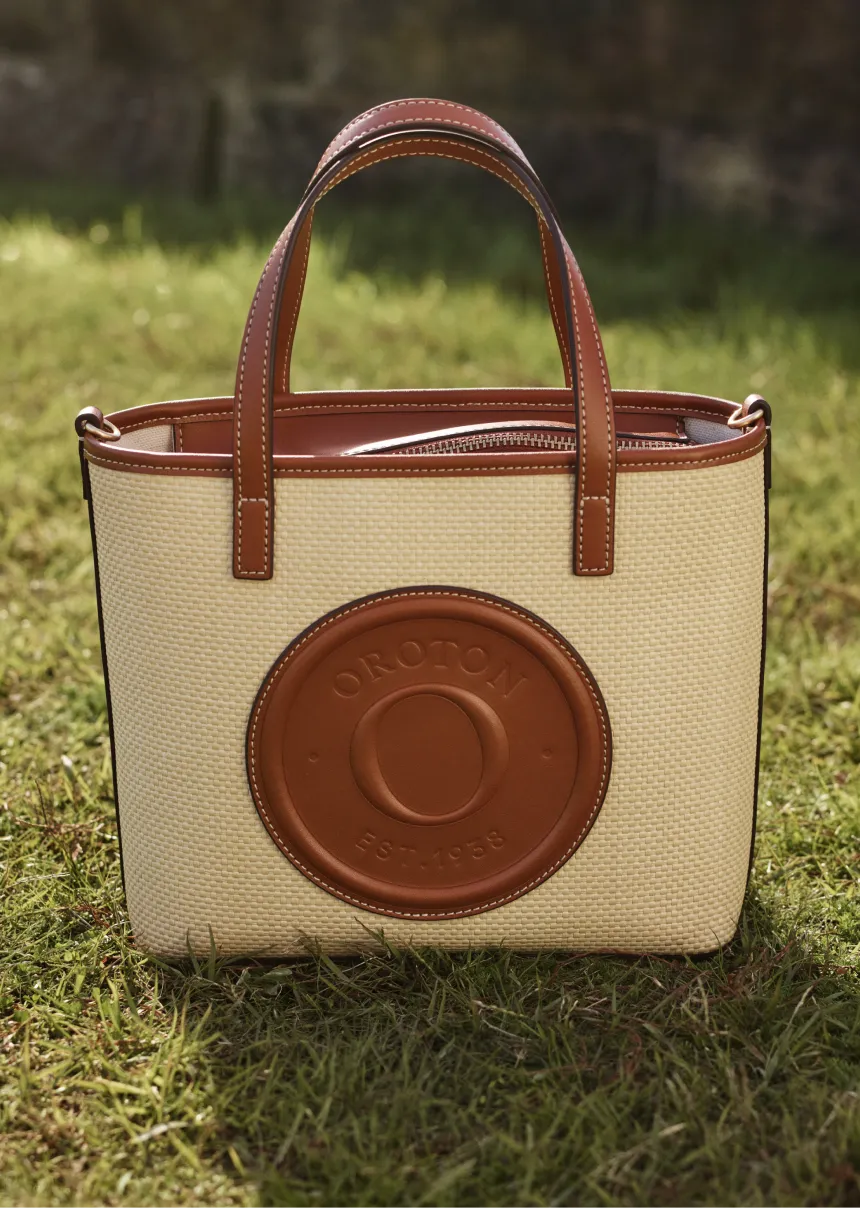 A stylish tote bag with a brown leather logo on a grassy background.