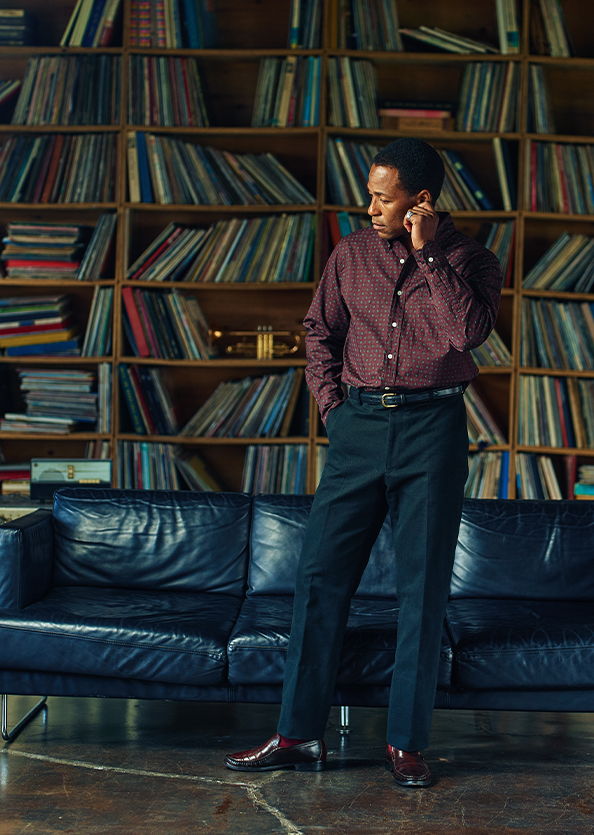 Person standing by a bookshelf, talking on the phone while wearing stylish clothes.