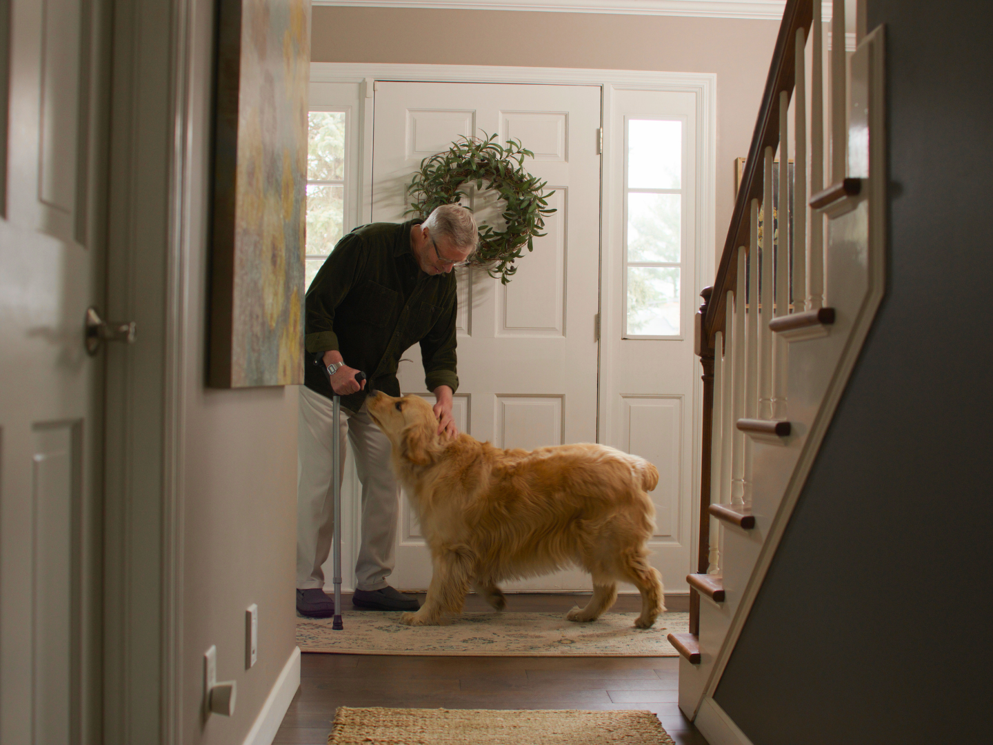 Older man using a Y-Cane while bending down to pet a golden retriever near a front door