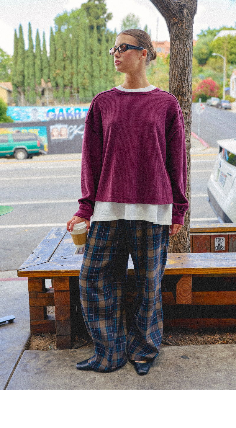 Person wearing a maroon sweater and plaid pants, holding a coffee cup outdoors.