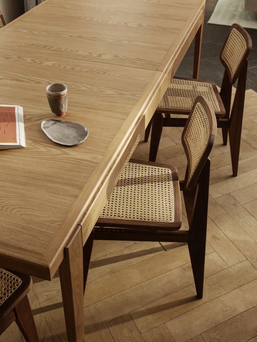 A wooden S-Extending dining table by Gubi with chairs, a coffee cup, and a magazine.