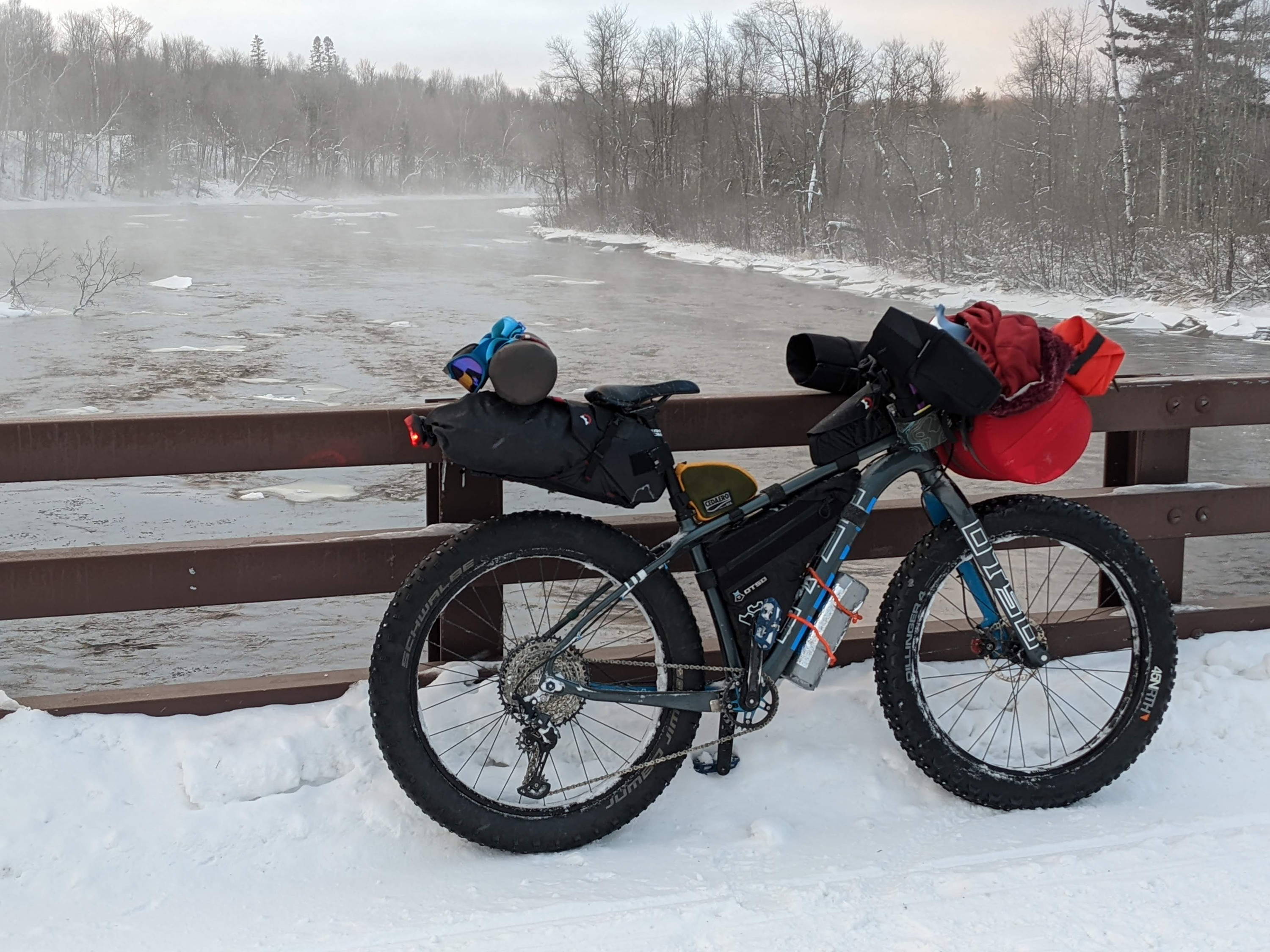 A fully-loaded Otso Voytek leans against a metal bridge railing while a winter river steams in the background.