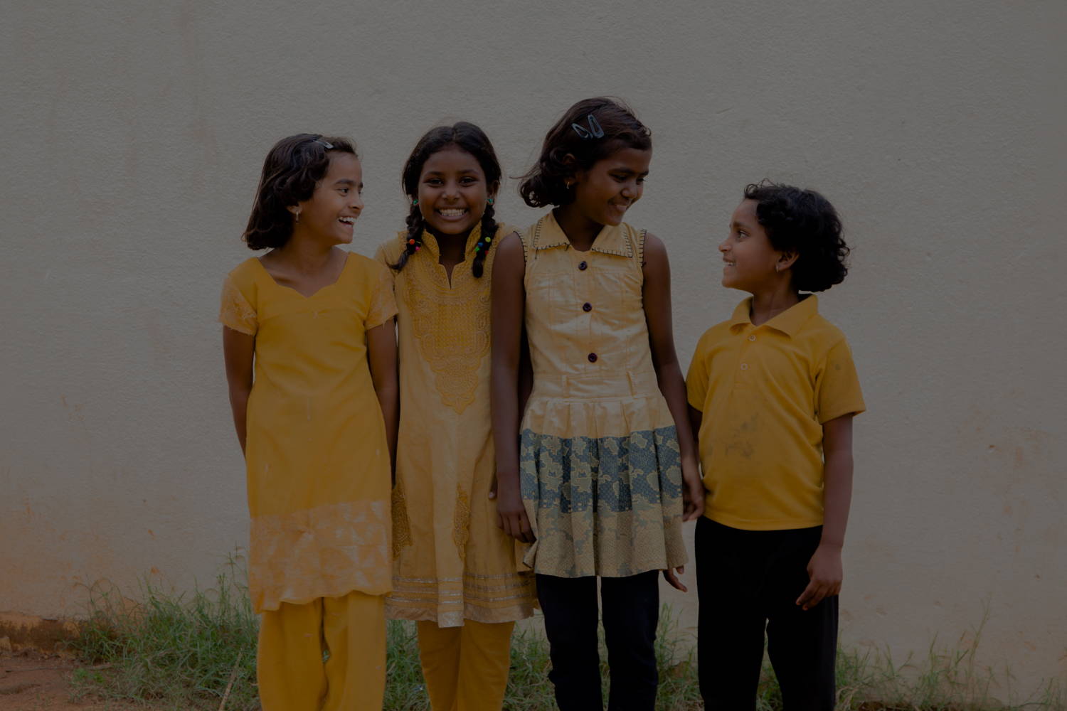 3 young Indian girls, hold hands while smiling, laughing, and looking at each other. They are all wearing yellow dresses.