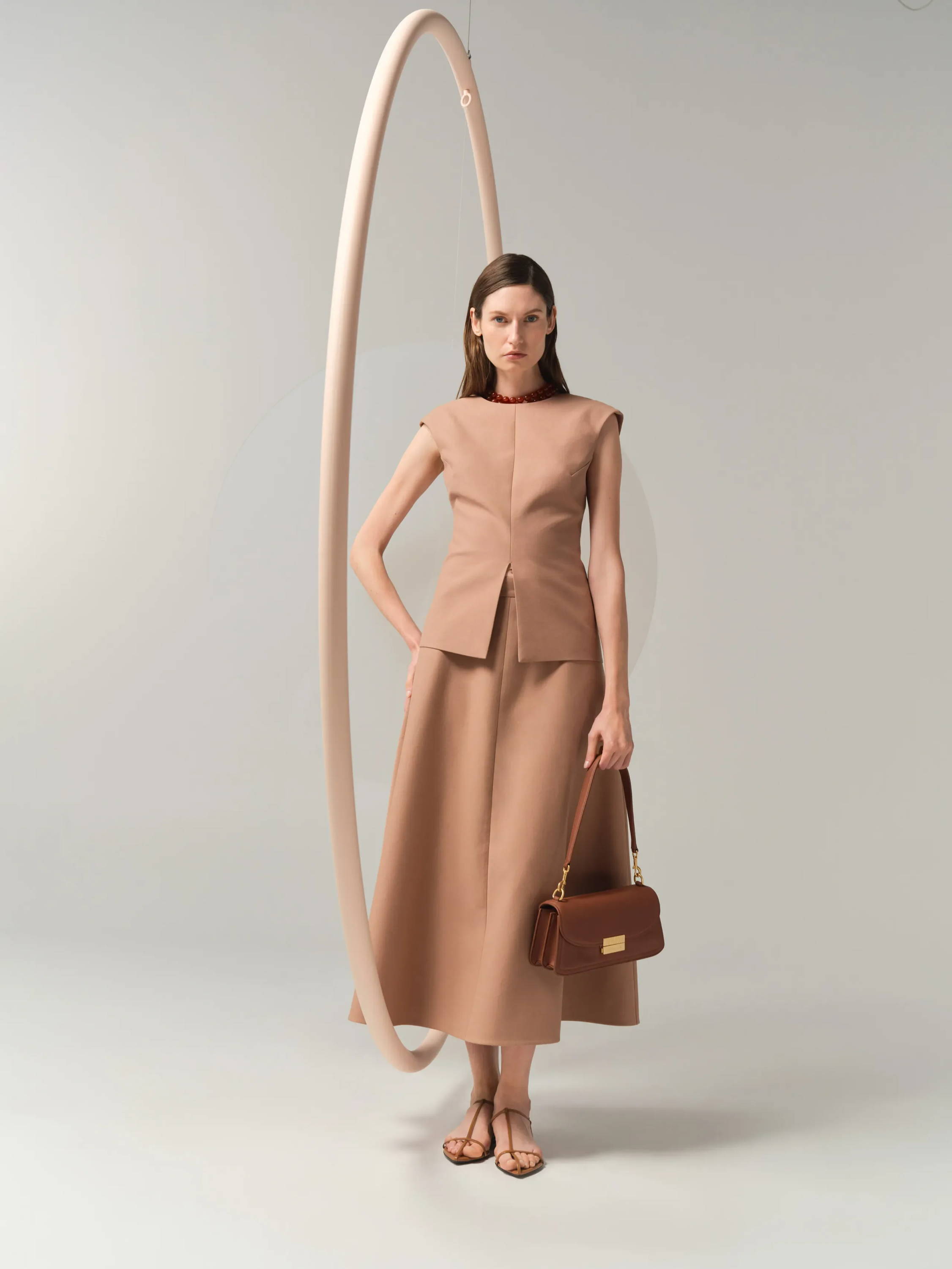 Model wearing a beige outfit with a handbag, standing near a circular backdrop.