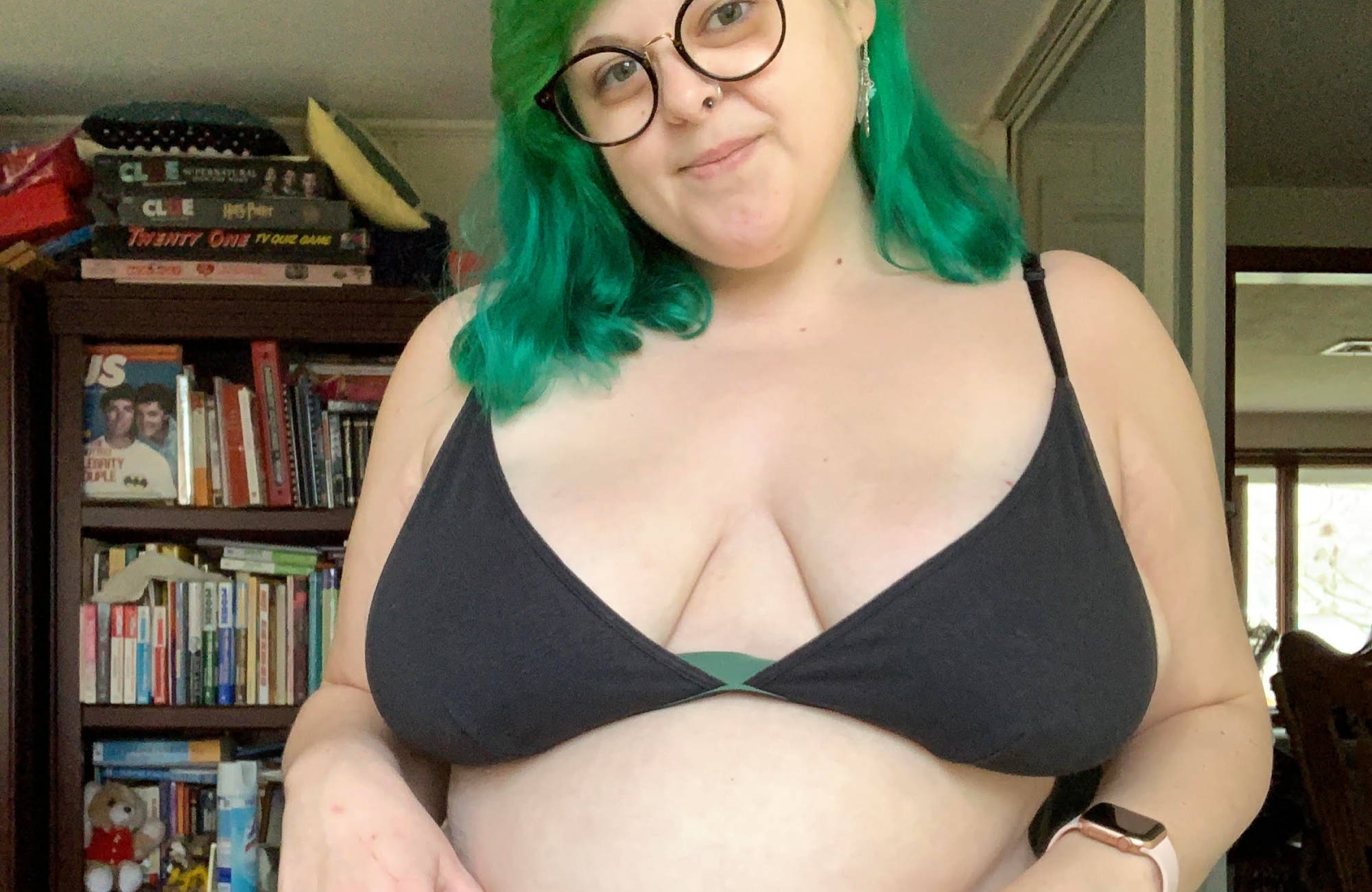 A woman with green hair and circle glasses in a black bralette stands in front of a bookshelf