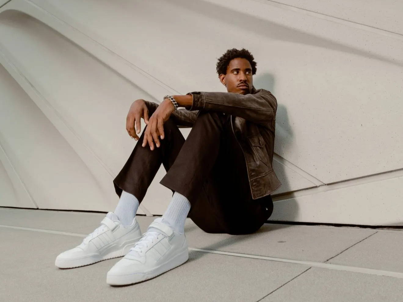 male model in white adidas forum shoes sitting against wall
