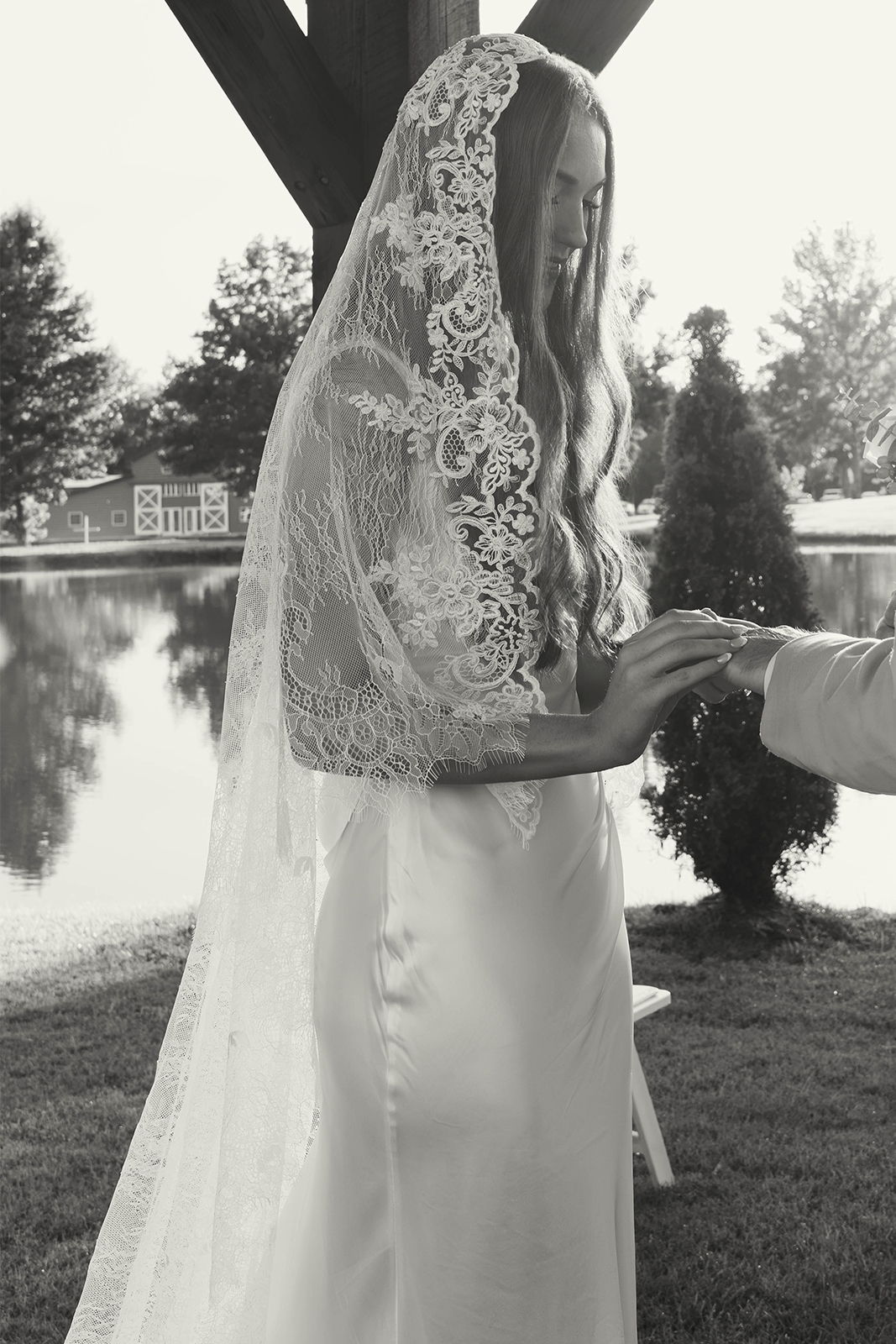 Bride in white dress and veil exchanging rings by a lake.
