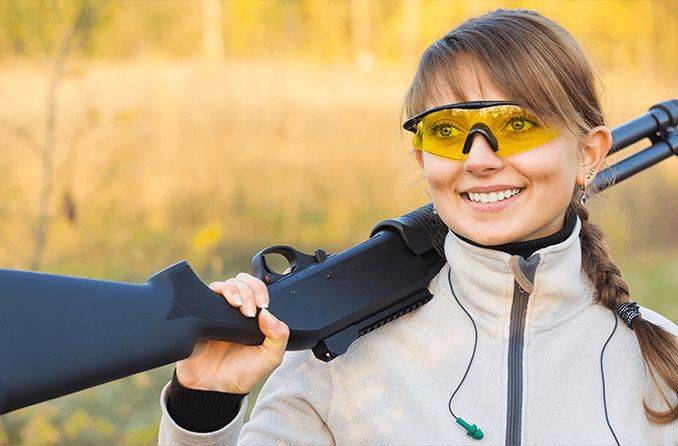 Une photo d'une femme portant des lunettes de nuit avec des verres teintés jaunes pour la chasse et tenant un fusil portant une veste beige avec une chemise noire