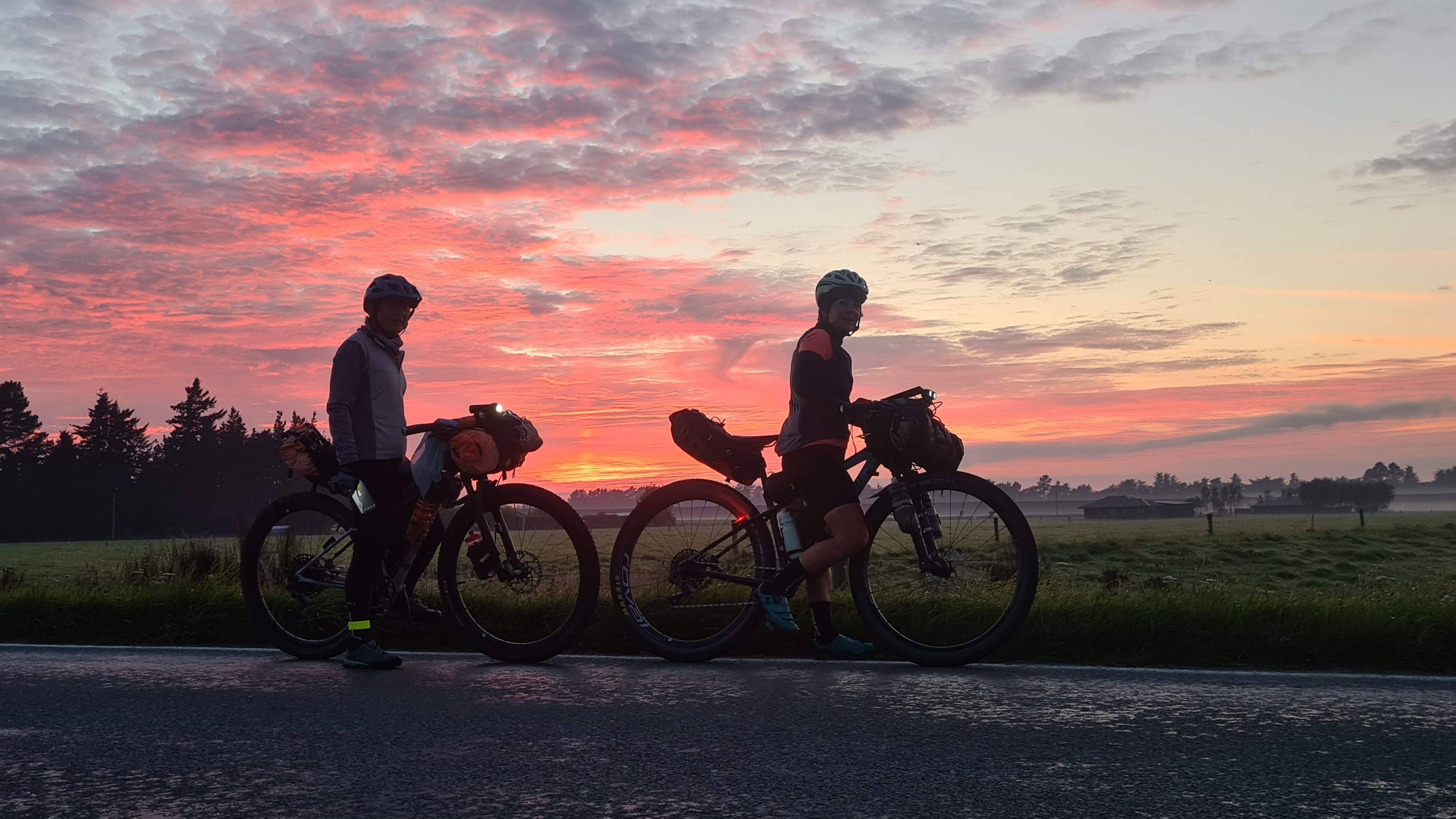 Two cyclists stand against a dramatic red sunset with their fully-loaded bikepacking rigs.