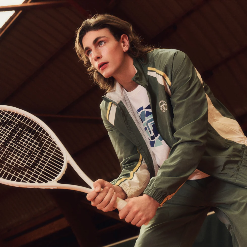 Young man holding a tennis racket, wearing a sporty Sergio Tacchini outfit indoors.