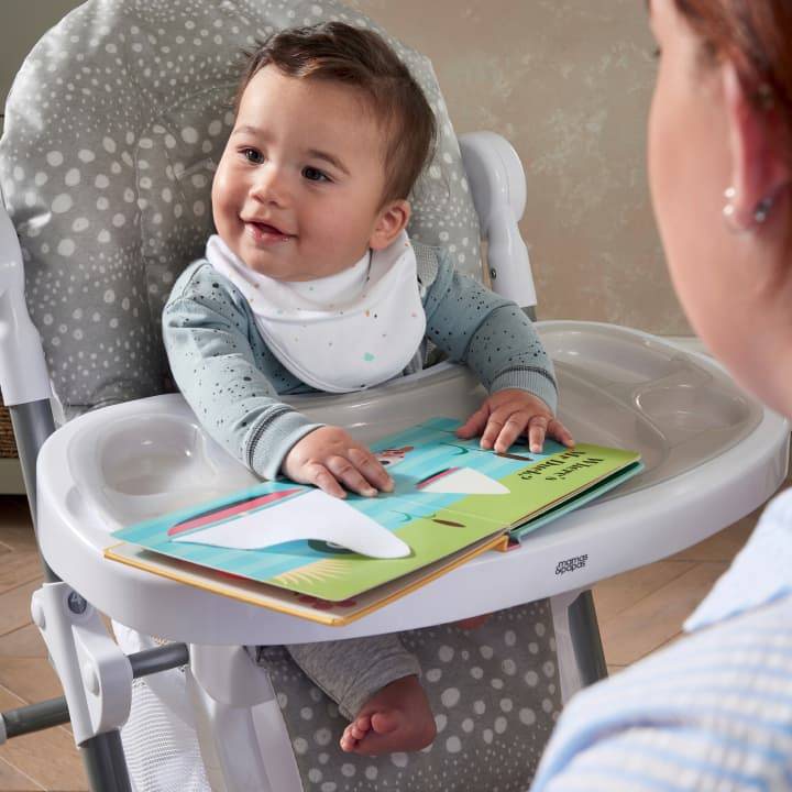 A little boy sits in the Mamas and Papas Snax high chair in grey, smiling up at someone beyond the camera.